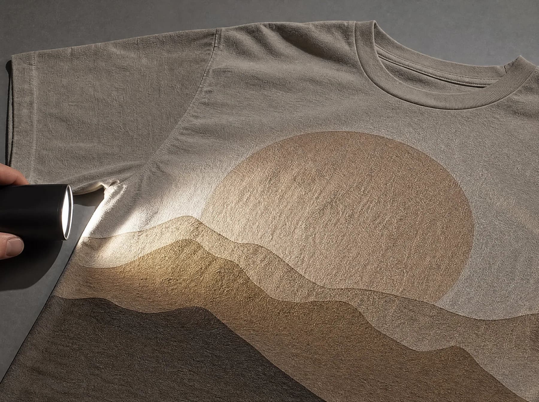 A printed t-shirt laid flat with a lamp held low at the side so the beam skims across the print.